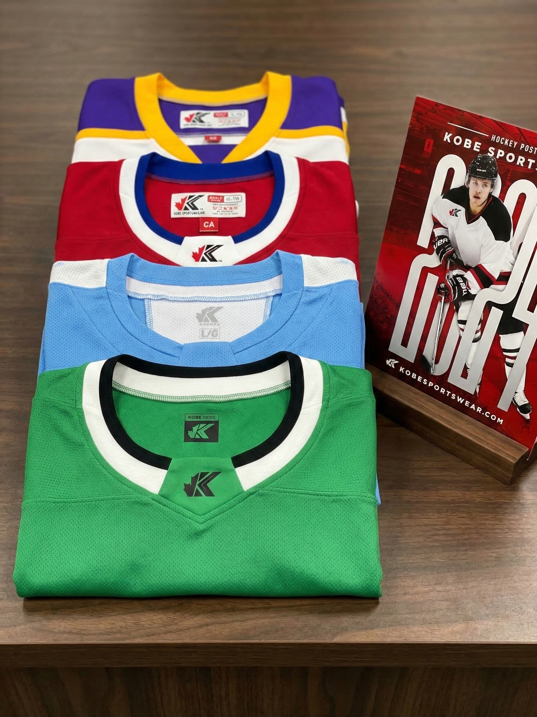 Four folded Kobe Sportswear jerseys in green, blue, red, and purple on a wooden surface with a book about Kobe sportswear memorabilia.